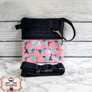 May include: Black crossbody bag with a pink panel featuring a cat print. The bag has a black adjustable strap and a zippered closure. The cat print includes cats in various colors wearing teal sweaters.