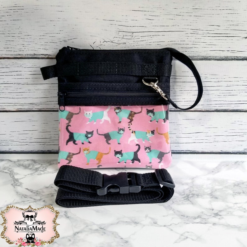 May include: Black crossbody bag with a pink panel featuring a cat print. The bag has a black adjustable strap and a zippered closure. The cat print includes cats in various colors wearing teal sweaters.