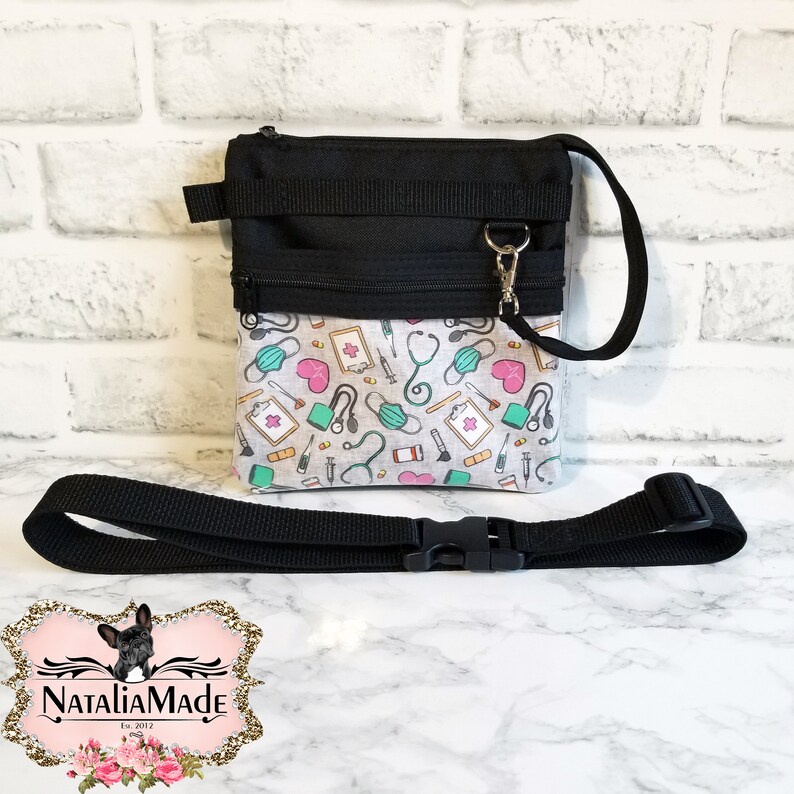 Op de afbeelding: Zwarte crossbodytas met een patroon aan de voorkant met medische symbolen zoals stethoscopen, harten en maskers. De tas heeft een zwarte band en een ritssluiting. Ontworpen voor medisch personeel.