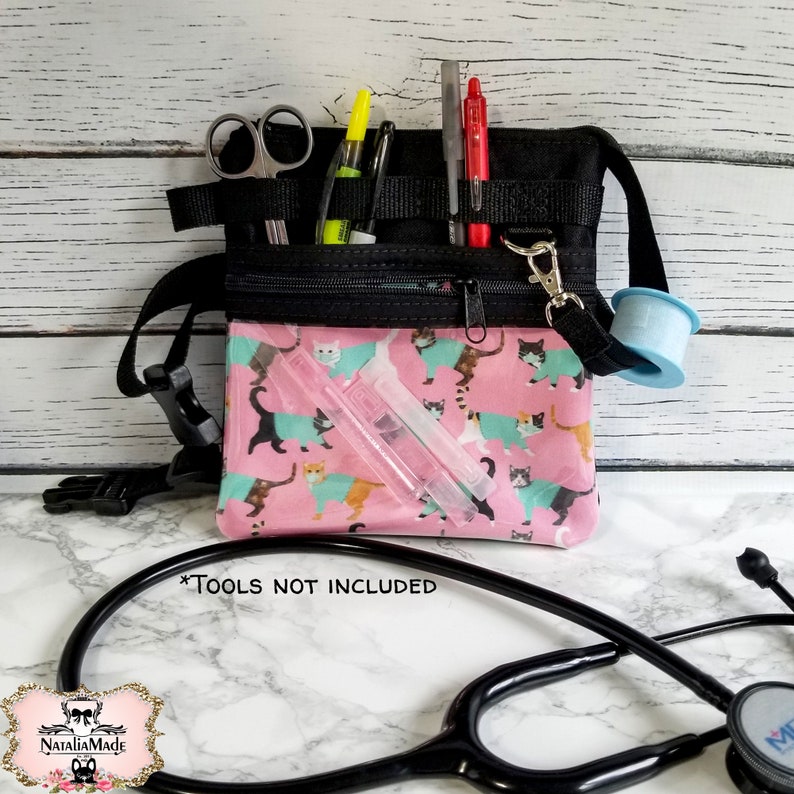 May include: Black and pink medical fanny pack with a cat print. The pack has multiple pockets and compartments for medical tools. The image includes scissors, pens, and tape. The text on the image says "TOOLS NOT INCLUDED".