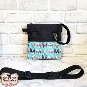 May include: Black crossbody bag with a teal panel printed with French bulldog faces wearing glasses. The bag has a black adjustable strap and a gold-coloured zip. The bag is designed to be worn across the body.