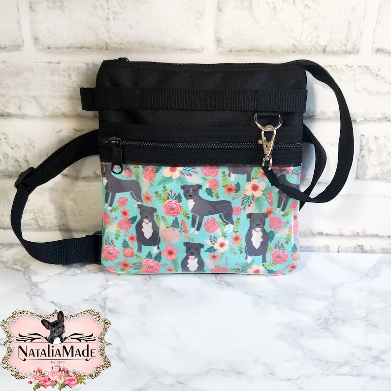 May include: Black and turquoise dog treat bag with a floral and dog pattern. The bag has a black strap and zipper closure. The bag has a silver clasp and the text "NataliaMade" on the bottom.