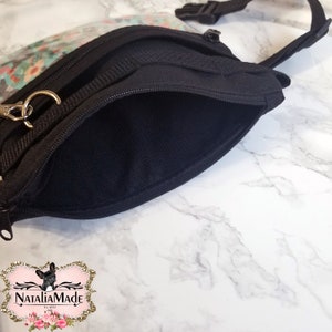 May include: Black fanny pack with a clear floral-patterned front pocket. The bag has a zippered main compartment and an adjustable strap with a buckle. A gold-colored metal clasp is attached to the bag. The bag is on a white marble surface.