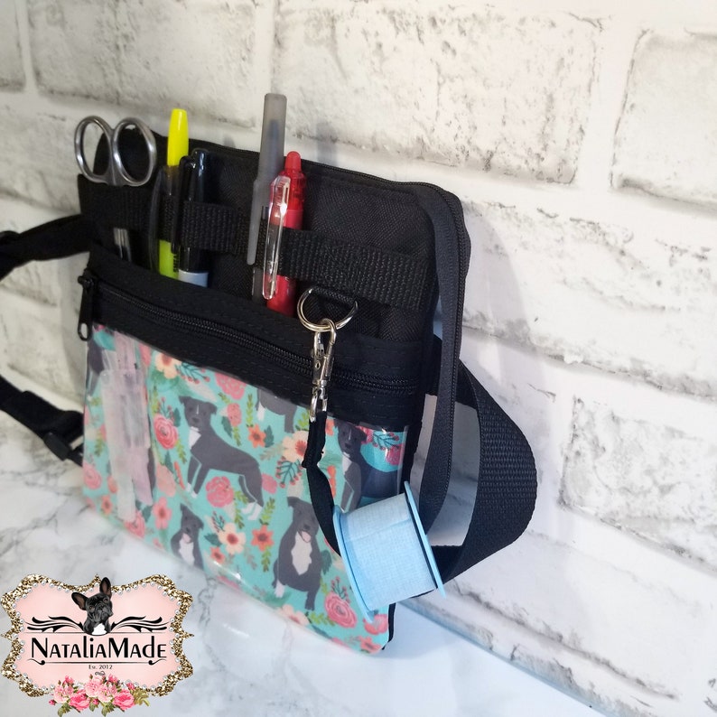 May include: Black and turquoise utility bag with a floral and dog pattern. The bag has a black strap, zipper, and pockets holding scissors, pens, and a spool of blue thread. The bag has a clear front pocket.