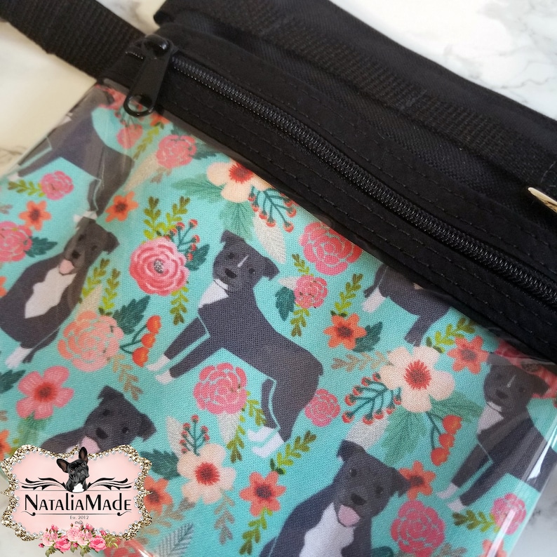 May include: A black zippered pouch with a teal floral and dog print. The pattern features gray dogs and pink flowers. The pouch has a black strap and zipper. The logo "NataliaMade" is visible in the bottom left corner.