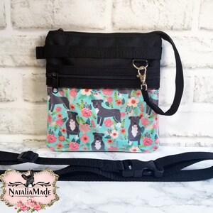 May include: Black crossbody bag with a floral pattern featuring pit bull illustrations. The bag has a black strap, zipper, and trim. The background is a light blue with pink and orange flowers.