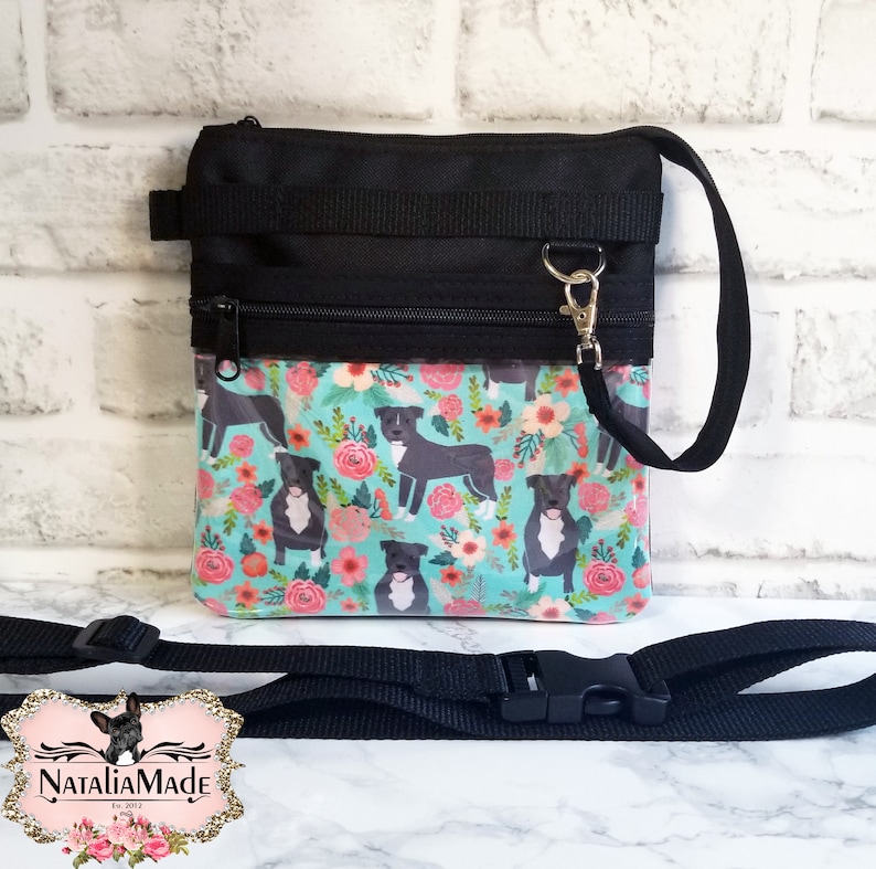 May include: Black crossbody bag with a floral pattern featuring pit bull illustrations. The bag has a black strap, zipper, and trim. The background is a light blue with pink and orange flowers.