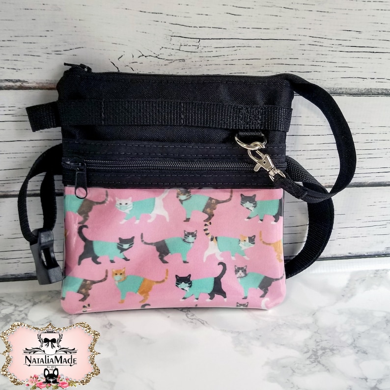May include: Black and pink crossbody bag with a cat pattern. The bag features a black top section with a zipper and strap attachments. The bottom section is pink with a repeating pattern of cats wearing teal sweaters and face masks.