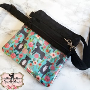 May include: Black crossbody bag with a clear front panel featuring a floral and dog pattern. The pattern includes black and white dogs and pink flowers on a teal background. The bag has a black adjustable strap and a zipper closure.