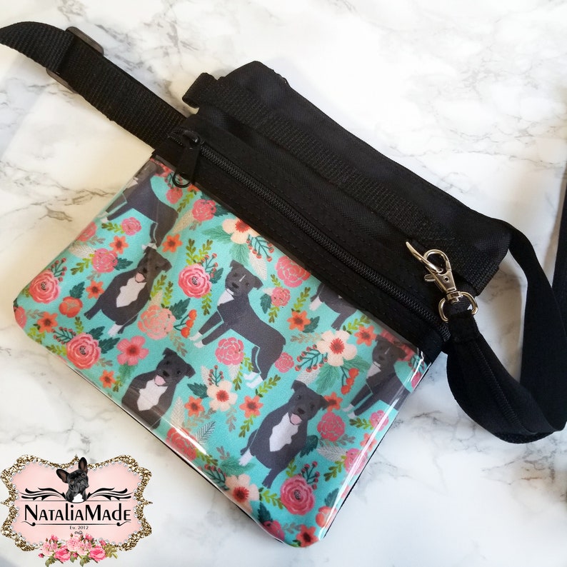 May include: Black crossbody bag with a clear front panel featuring a floral and dog pattern. The pattern includes black and white dogs and pink flowers on a teal background. The bag has a black adjustable strap and a zipper closure.