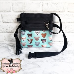 May include: Black and teal crossbody bag with a dog-themed pattern. The bag features a black adjustable strap, a zippered main compartment, and a smaller front pocket. The teal fabric is printed with various dog faces wearing glasses.
