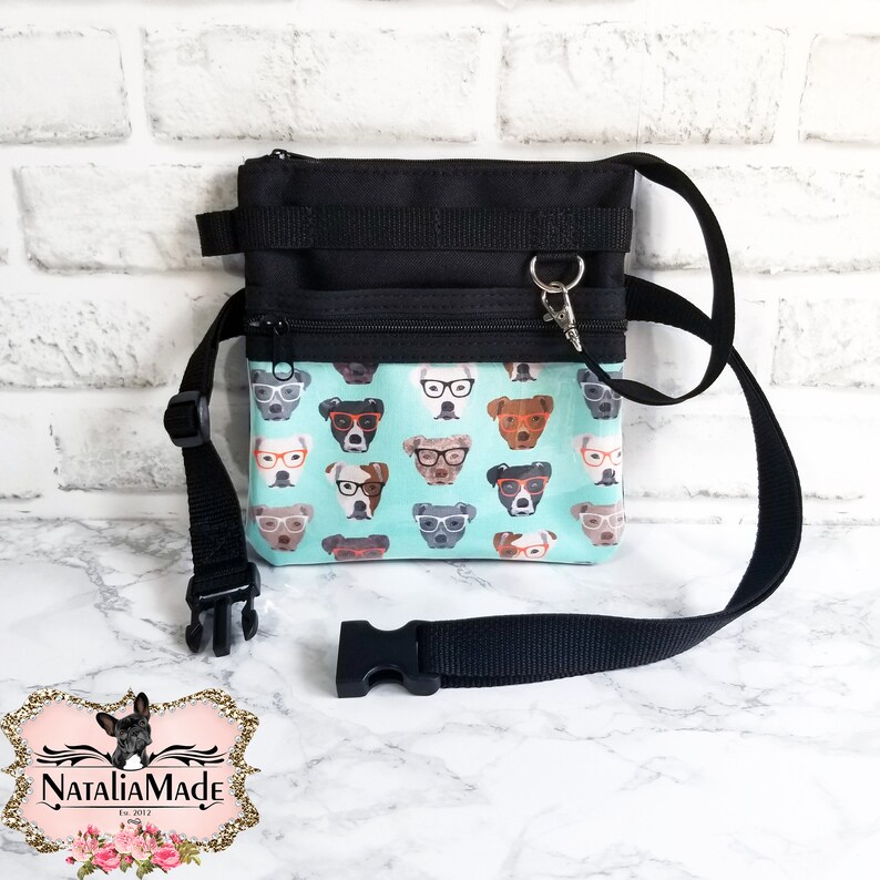 May include: Black and teal crossbody bag with a dog-themed pattern. The bag features a black adjustable strap, a zippered main compartment, and a smaller front pocket. The teal fabric is printed with various dog faces wearing glasses.