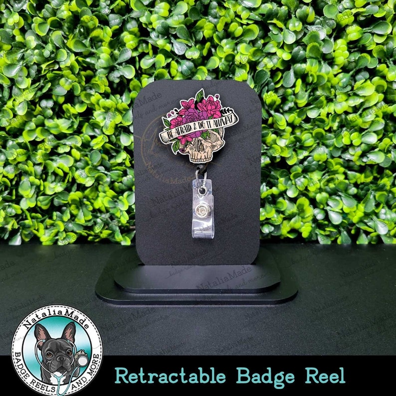 May include: Retractable badge reel with a black skull and pink flowers. The skull has a banner that reads "Be Afraid & Do It Anyway".