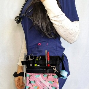 May include: A navy blue medical uniform with a black utility belt. The belt has multiple pockets holding pens, scissors, and other medical tools. A pink pouch with a cat pattern is attached. A stethoscope hangs around the neck.