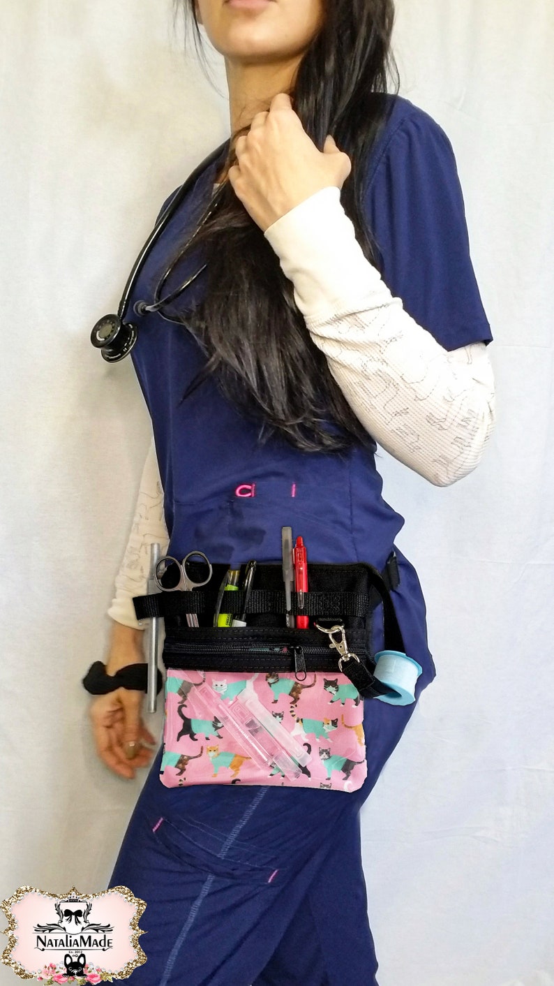 May include: A navy blue medical uniform with a black utility belt. The belt has multiple pockets holding pens, scissors, and other medical tools. A pink pouch with a cat pattern is attached. A stethoscope hangs around the neck.