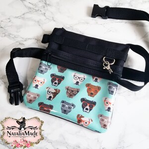 May include: A black and teal dog treat bag with a repeating pattern of dogs wearing glasses. The bag has a black adjustable strap and a zippered pocket. The bag is made by NataliaMade.