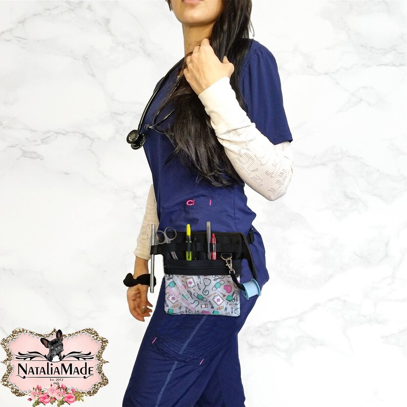 Op de afbeelding: Een zwarte medische gereedschapsriem met een patroonvak met stethoscopen, harten en medische symbolen. De riem bevat pennen, scharen en ander medisch gereedschap. Het model draagt een marineblauwe broek en een stethoscoop.