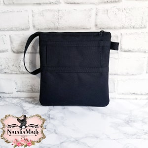 May include: A black crossbody bag with a zippered top and a small handle. The bag has two horizontal stitched lines across the front. The bag is sitting on a white and gray marble surface.