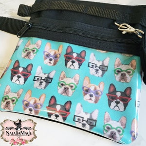 May include: Black crossbody bag with a teal front panel featuring a repeating pattern of French bulldogs wearing glasses in various colours. The bag has a black zip, strap, and a silver-coloured clasp. The NataliaMade logo is in the bottom left corner.