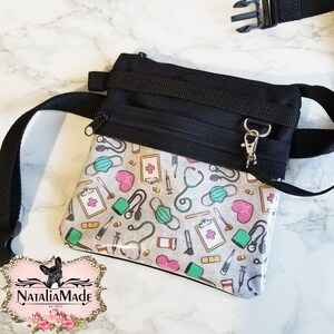 Op de afbeelding: Zwarte heuptas met een medisch thema patroon, inclusief stethoscopen, gezichtsmaskers, harten en medische kaarten. De tas heeft een zwarte verstelbare riem en een zilveren sluiting.
