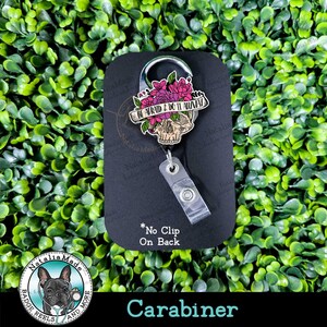 May include: A black and silver carabiner with a skull and floral design. The skull has a pink and white floral crown and a banner that reads "BE AFRAID & DO IT ANYWAY". The carabiner has a clear plastic badge reel attached to it.
