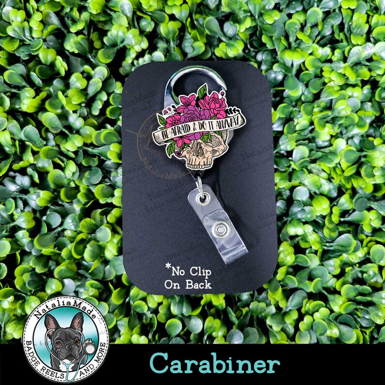 May include: A black and silver carabiner with a skull and floral design. The skull has a pink and white floral crown and a banner that reads "BE AFRAID & DO IT ANYWAY". The carabiner has a clear plastic badge reel attached to it.