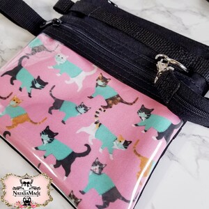 May include: A small, pink zippered pouch with a repeating pattern of cats wearing teal sweaters and face masks. The pouch has a clear front, black zipper, and black strap. The brand logo is in the bottom left corner.