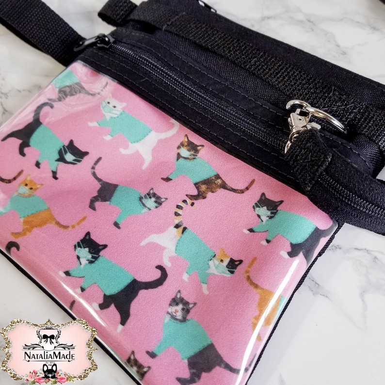 May include: A small, pink zippered pouch with a repeating pattern of cats wearing teal sweaters and face masks. The pouch has a clear front, black zipper, and black strap. The brand logo is in the bottom left corner.
