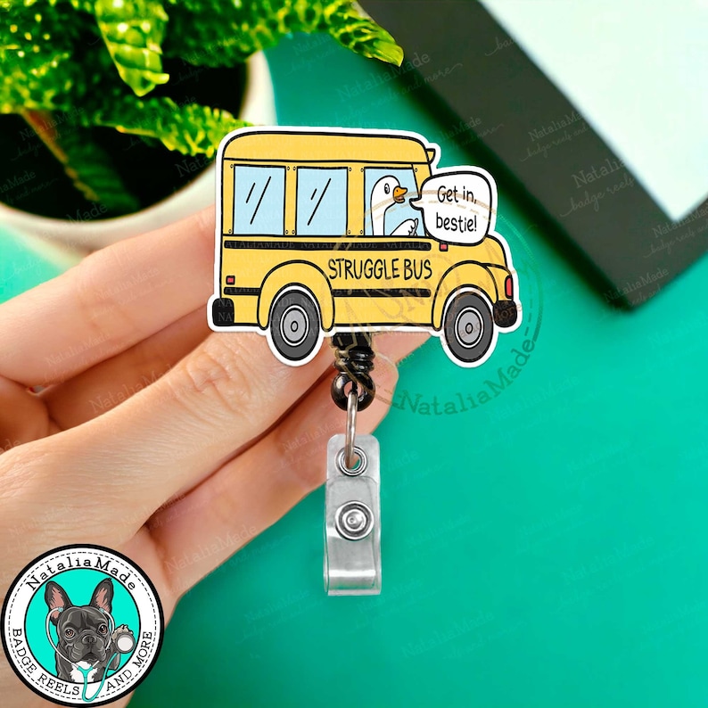 May include: A yellow school bus badge reel with the text "STRUGGLE BUS" and a cartoon duck. The duck has a speech bubble that says "Get in, bestie!". The badge reel has a black retractable cord and a silver clip.