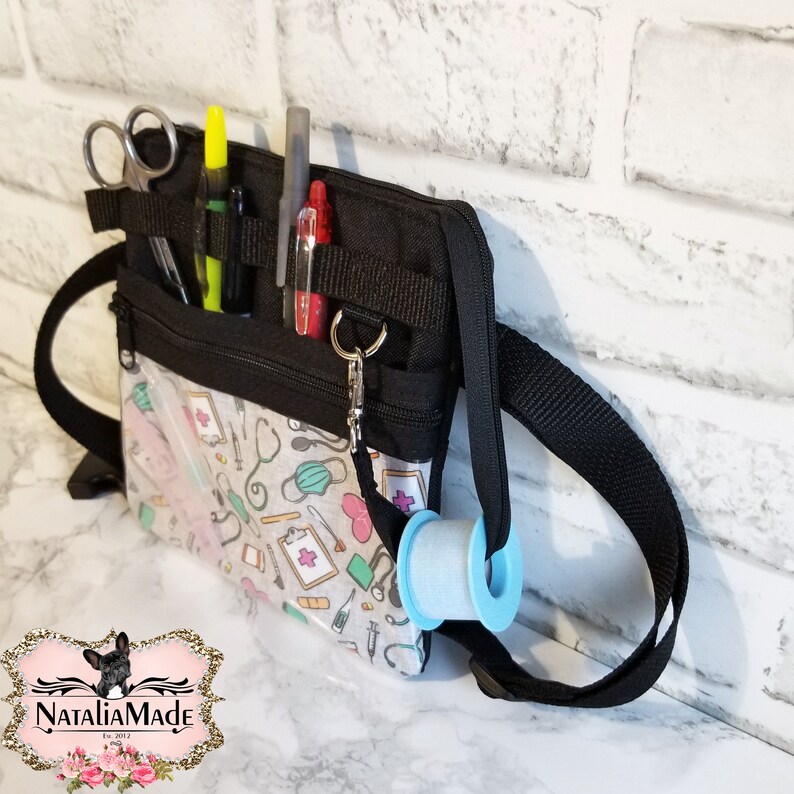 Op de afbeelding: Zwarte medische utility-tas met een doorzichtige voorzak met een medisch thema. De tas heeft meerdere compartimenten met een schaar, pennen en een rol tape. Een zwarte schouderriem is bevestigd.