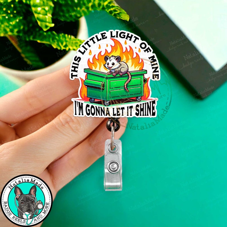 May include: A green trash can with flames and a cartoon possum inside. The text on the badge reel reads "This little light of mine I'm gonna let it shine".