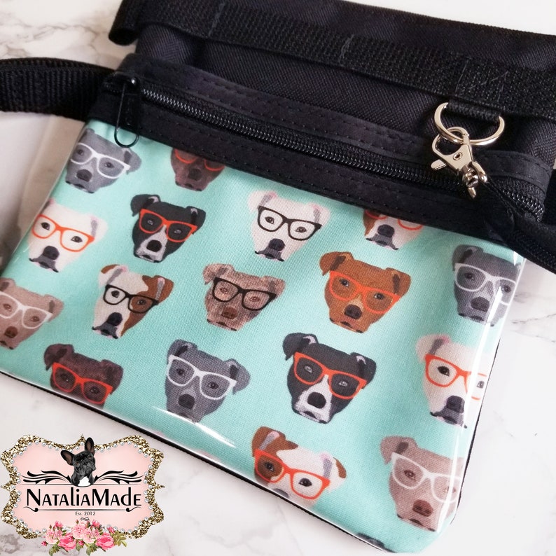 May include: A black and teal dog treat bag with a zipper closure and a silver clasp. The teal front features a repeating pattern of dogs wearing glasses. The bag has a black strap and a logo in the bottom left corner.
