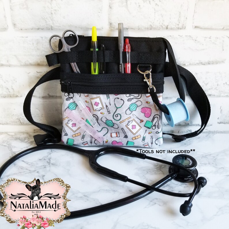 Op de afbeelding: Zwarte medische heuptas met een patroon, met een zwarte riem, met pennen, schaar en medische benodigdheden. De tas heeft een rits en een patroon van medische iconen. Een stethoscoop staat op de voorgrond.