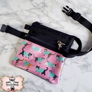 May include: A black fanny pack with a pink front panel featuring a cat pattern. The cats wear teal and orange sweaters. The bag has a black adjustable strap and a silver-tone zipper pull.
