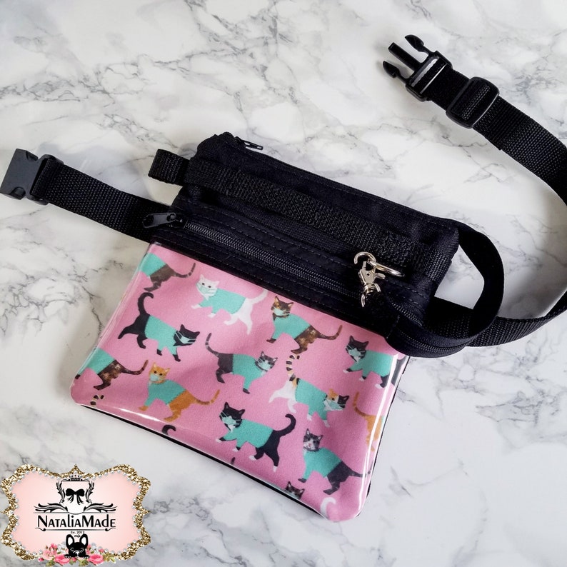 May include: A black fanny pack with a pink front panel featuring a cat pattern. The cats wear teal and orange sweaters. The bag has a black adjustable strap and a silver-tone zipper pull.