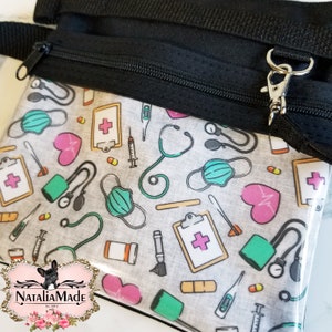 Op de afbeelding: Een doorzichtige etui met ritssluiting, zwarte band en zilveren sluiting. De etui heeft een herhalend patroon van medische symbolen, waaronder stethoscopen, maskers en harten, op een grijze achtergrond. De merknaam "NataliaMade" is linksonder zichtbaar.