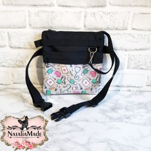 Op de afbeelding: Zwarte en witte crossbody tas met een medisch thema patroon. De tas heeft een zwart bovenstuk met een rits en een patroon onderstuk met stethoscopen, maskers en medische symbolen. Het heeft een verstelbare zwarte riem en een zilveren sluiting.