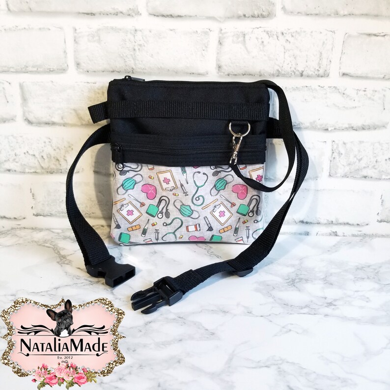 Op de afbeelding: Zwarte en witte crossbody tas met een medisch thema patroon. De tas heeft een zwart bovenstuk met een rits en een patroon onderstuk met stethoscopen, maskers en medische symbolen. Het heeft een verstelbare zwarte riem en een zilveren sluiting.