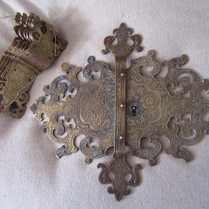 May include: Two ornate brass pieces with intricate floral designs. The larger piece is a decorative hinge with a keyhole, while the smaller piece is a hinged clasp with multiple folds.