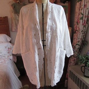 May include: White cotton jacket with a lace design and two pockets. The jacket is open in the front and has a button closure.
