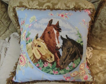 May include: A blue pillow with a brown fringe border features a circular embroidered design of three horses in a field of flowers. The horses are brown, white, and black.