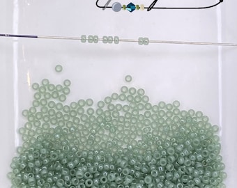 May include: A large pile of light green seed beads on a white surface. The beads are small and round, and they are arranged in a random pattern. There is a needle with a few beads threaded on it in the top of the image.