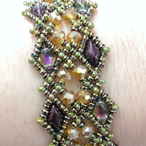 Op de afbeelding: Een kralenarmband met een geweven ontwerp met gouden, groene en paarse kralen. De armband zit om een pols.