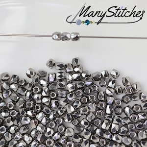 May include: A close-up of a pile of silver-colored faceted beads. The beads are small and have a shiny finish. They are scattered on a white surface.