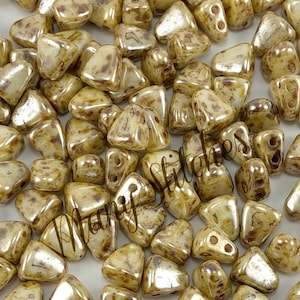 May include: A close-up of a pile of small, triangular, beige and brown glass beads with a metallic sheen. The beads have two holes each and are scattered on a white surface.