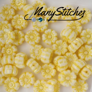 May include: A close-up of a pile of white glass flower beads with yellow accents. The beads are small and have a raised flower design.
