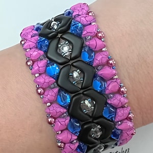 May include: A pink and blue beaded bracelet with black hexagonal beads and silver accents. The bracelet is on a person's wrist. The text "Many Stitches" is visible on the bracelet.