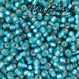 May include: A close-up image of a large quantity of teal blue seed beads. The beads are opaque and have a slightly frosted finish. The beads are arranged in a random pattern, creating a textured surface.