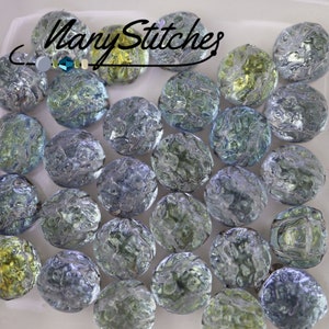May include: A collection of iridescent glass beads in shades of blue, green, and silver. The beads have a textured, organic appearance and are scattered across a white surface. The text "Many Stitches" is visible in the upper left corner.
