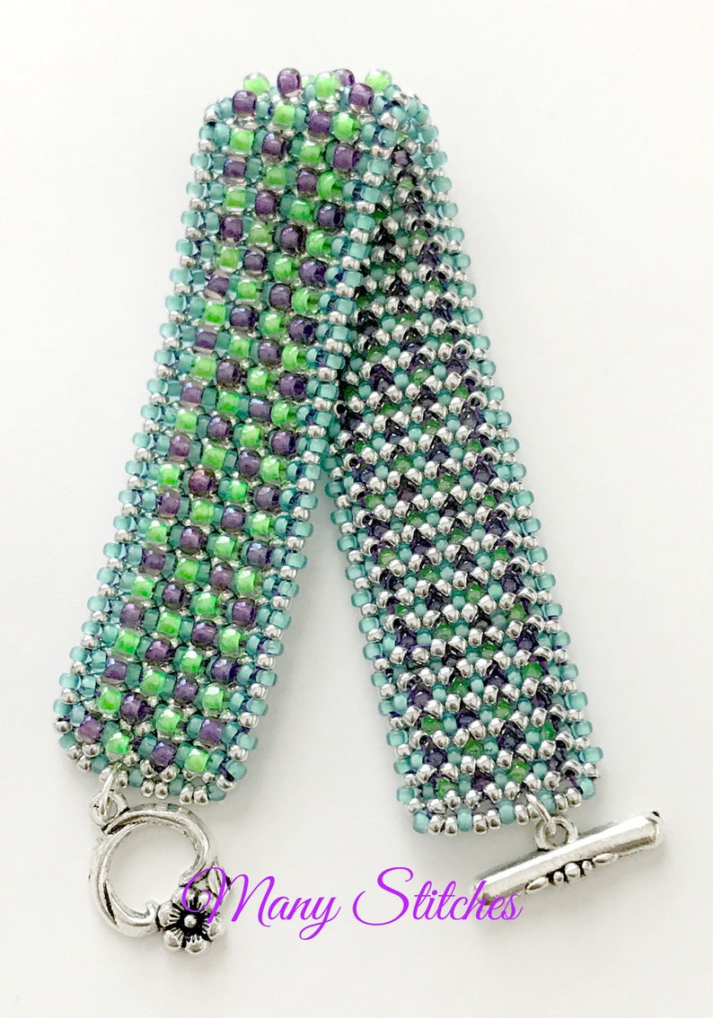 K&ouml;nnte beinhalten: Ein gr&uuml;nes, blaues und silbernes Perlenarmband mit einem Schiebeverschluss. Das Armband ist aus kleinen Perlen gefertigt und hat einen dezenten Ombre-Effekt. Das Armband ist mit "Mary Stitches" signiert.